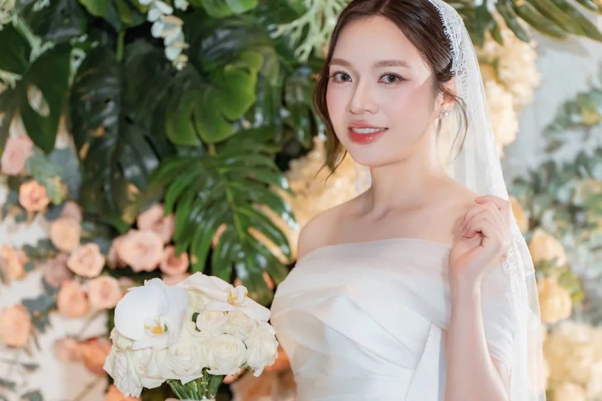 Cô dâu váy cưới trắng trễ vai cầm bó hoa trắng giữa phông hoa