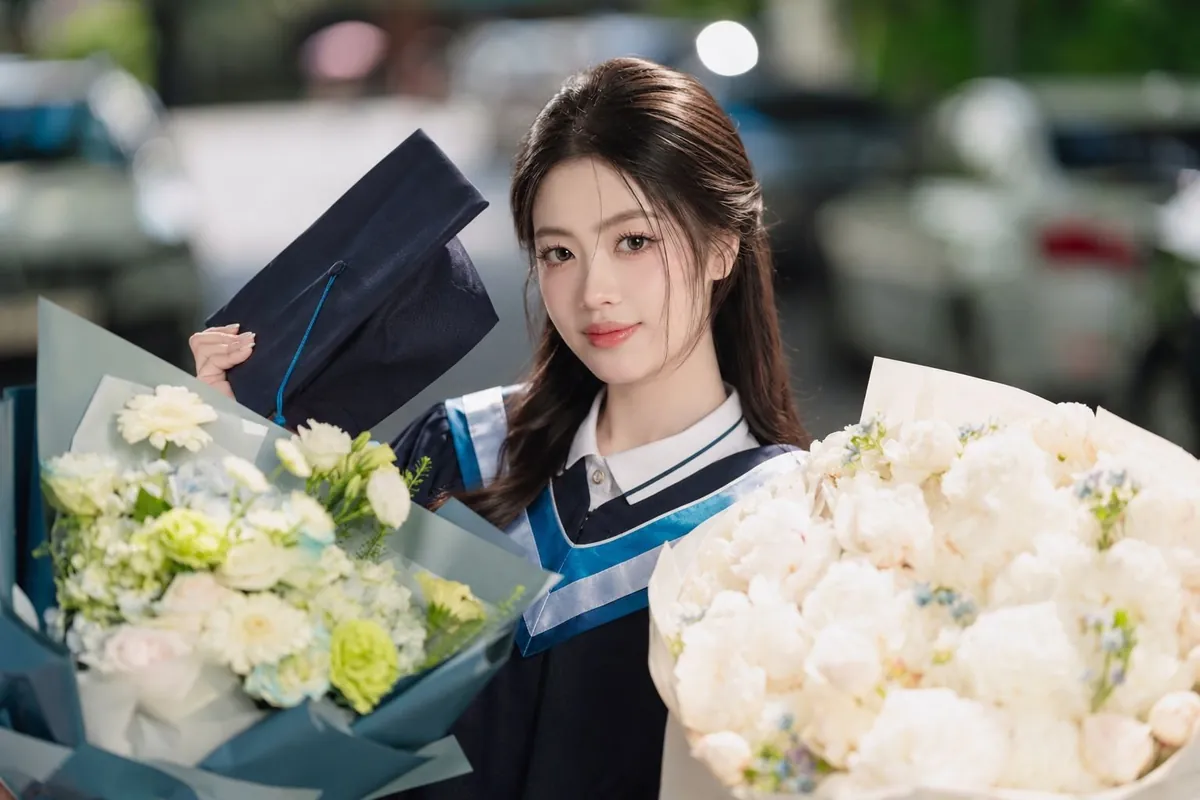 Nữ sinh mặc áo cử nhân xanh, ôm hai bó hoa trắng, tay cầm mũ tốt nghiệp