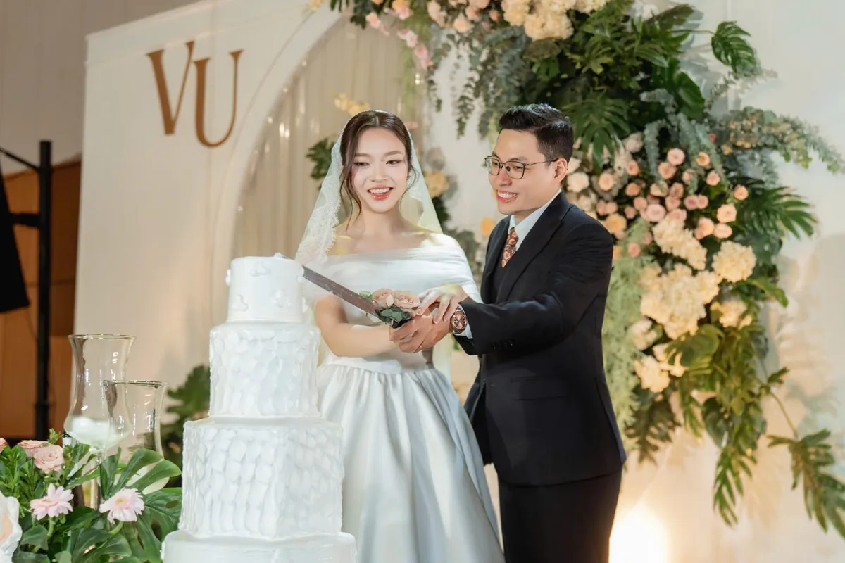 Cô dâu chú rể đứng bên bánh cưới nhiều tầng giữa hoa tươi